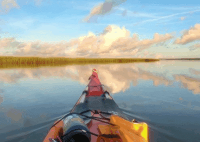 The view of over the helm of a kayak on Shem Creek with glassy water and the sky reflected in the sea.